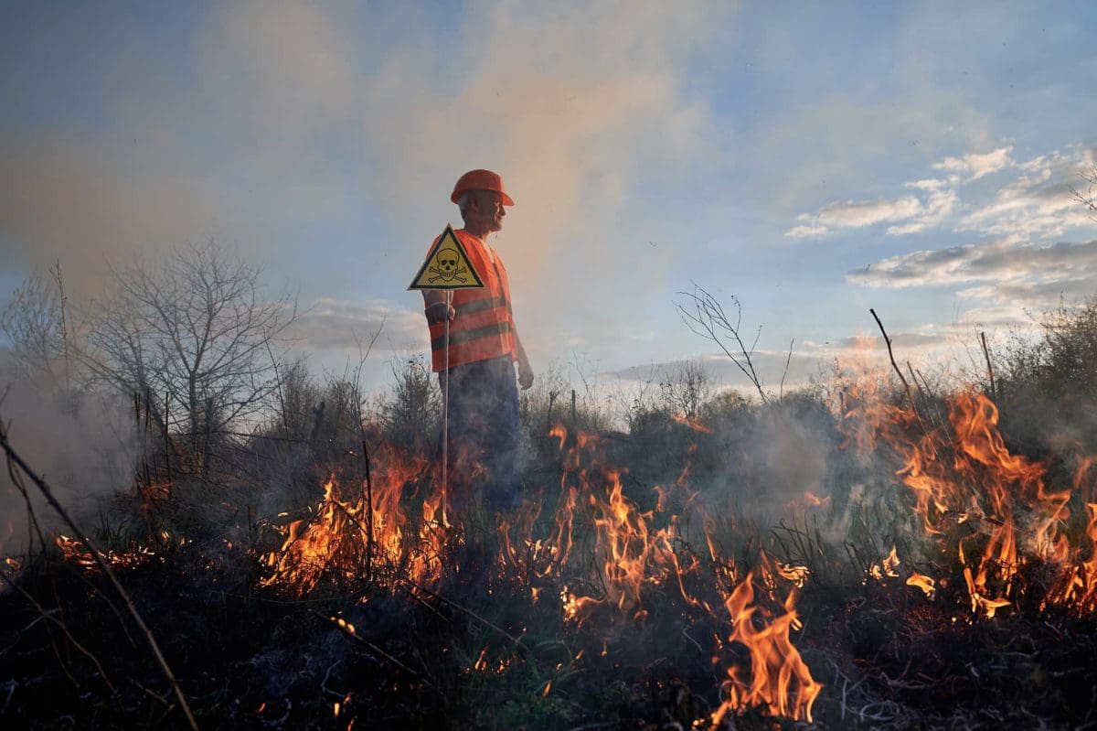 Fireman ecologist fighting fire in field in evening. Man in vest and helmet near burning grass with smoke, holding yellow triangle with skull and crossbones warning sign. Natural disaster concept.