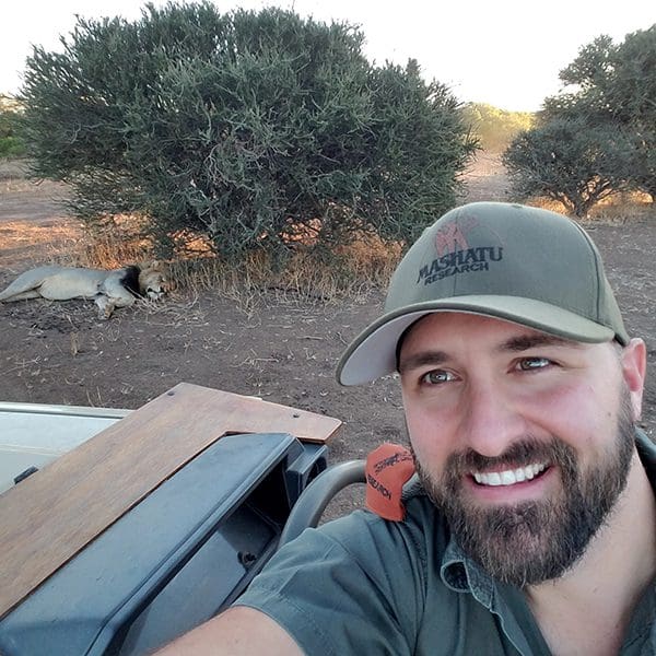 Dr. Snyman smiles at the camera in a sage green ball cap as a lion sleeps under a bush behind him.