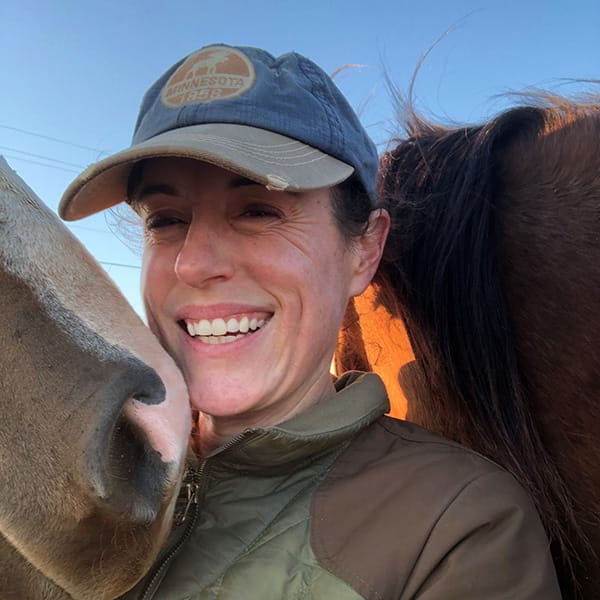 A woman with brown hair in a blue baseball cap smiles as two brown horses nuzzle her.