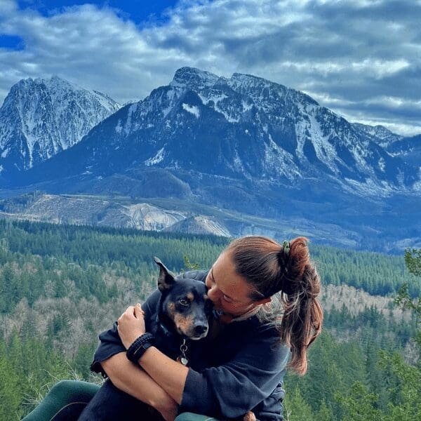 A young person smiles at a dog with a snow capped mountain in the background.