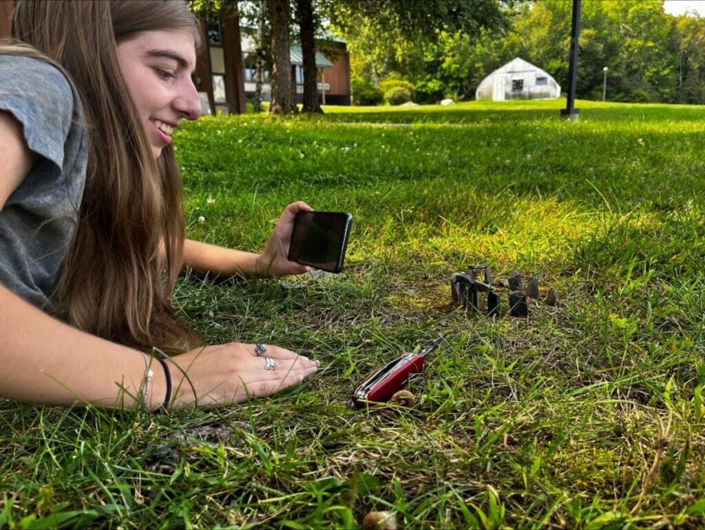 A younger person with a camera smiles at a tiny recreation of Stonehenge in the grass.