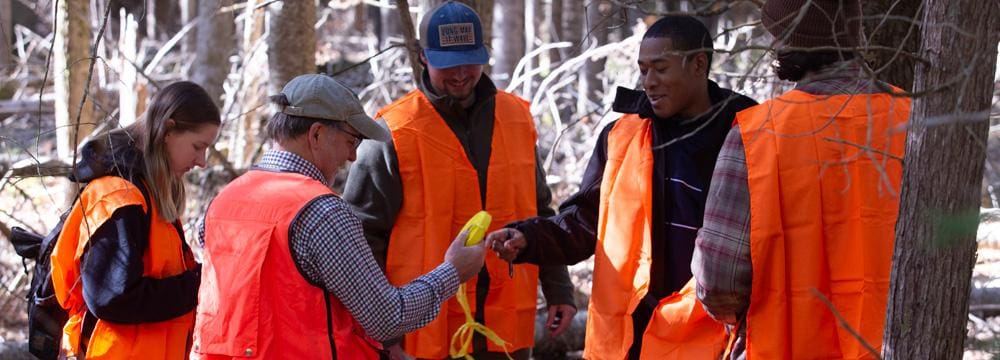 A group of students in bright hunting orange vests examine a yellow monitor in the woods.