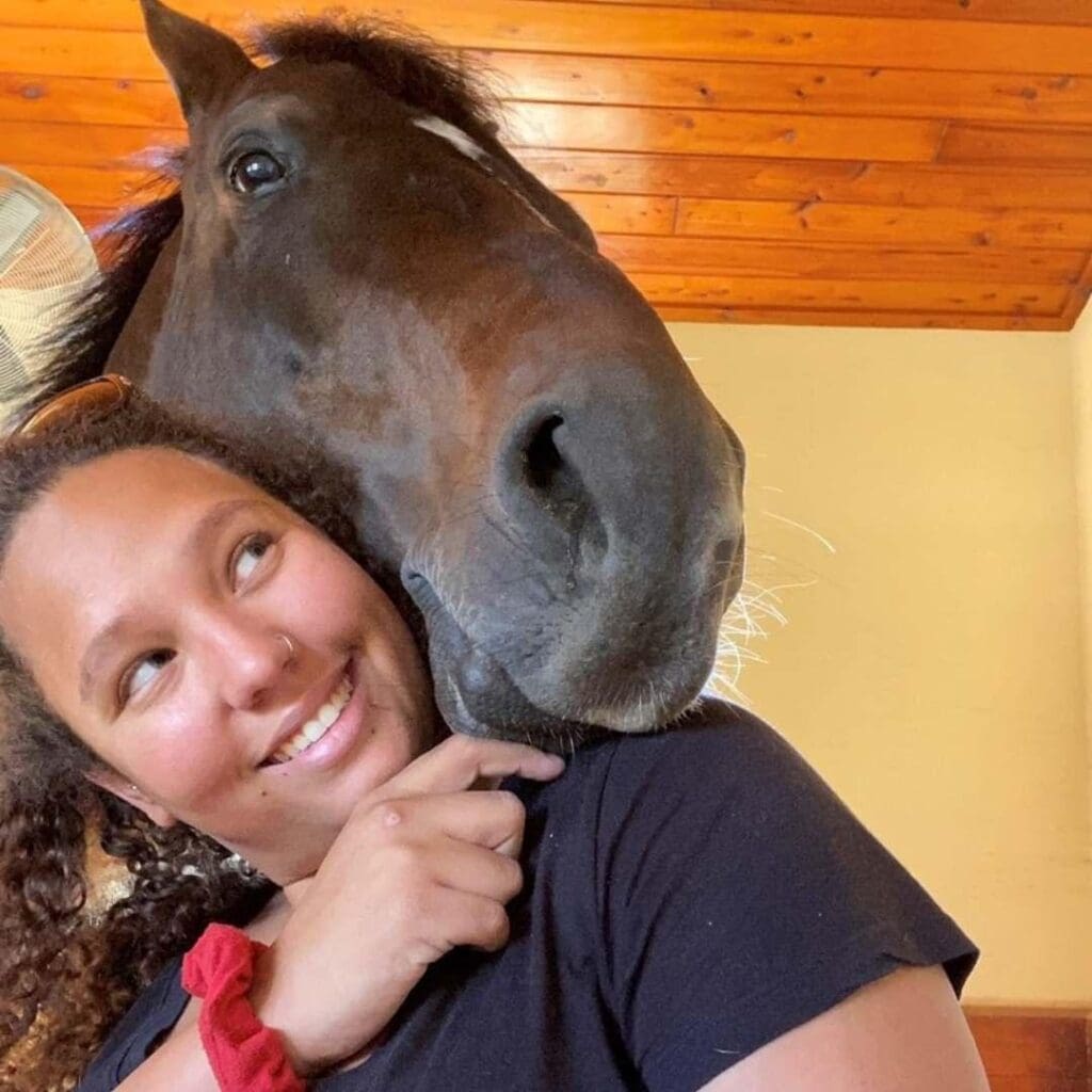 A young person with a bright red hair scrunchie smiles with their horse.