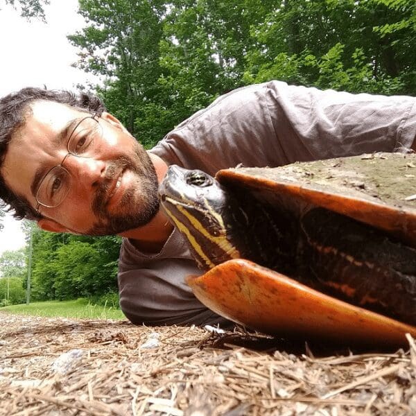 A young person with a beard lays on the ground next to a red bellied slider.