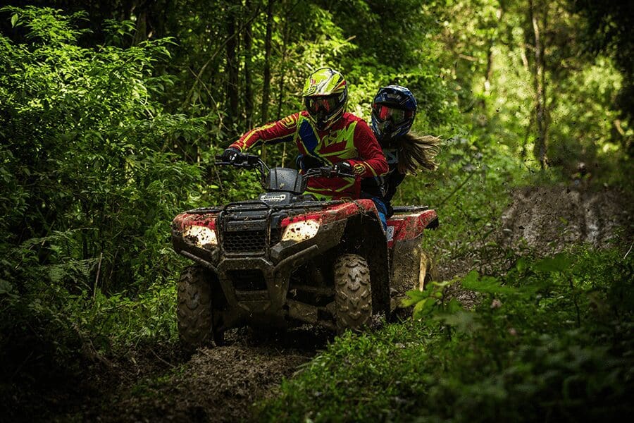 Two people clad in colorful gear and helmets speed through a muddy trail on a four-wheeler.