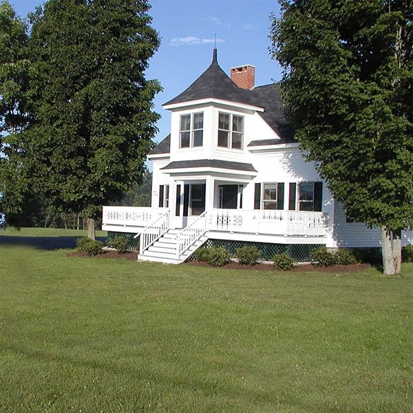 A classic farmhouse painted white with a black roof and brick chimney sits in a grassy field on a sunny day.