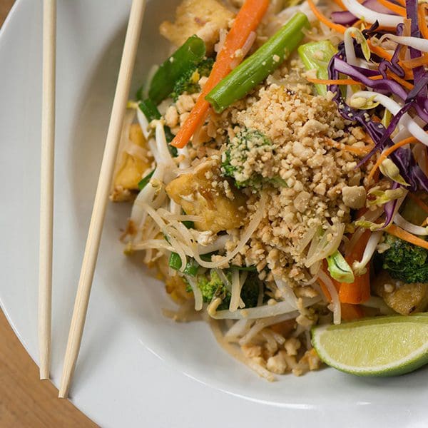 A pair of chopsticks rest on the side of a white bowl that contains a vegan pad Thai serving.