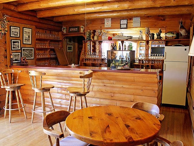 A wood-lined kitchen and bar area with tables and chairs gleams with bottles and glasses along the walls.