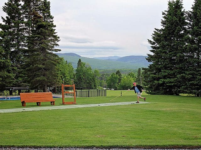A person in a red hat and jacket plays shuffleboard in a field near a pool in front of some small mountains.