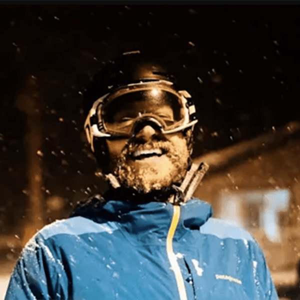 A person with a beard, black helmet, goggles, and blue snow jacket laughs during a night ski event.