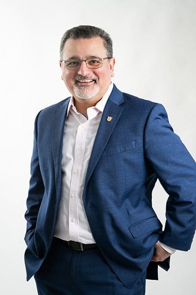 Dr. Melik Peter Khoury smiles at the camera in front of a white background in a dark blue suit coat and white collared shirt.