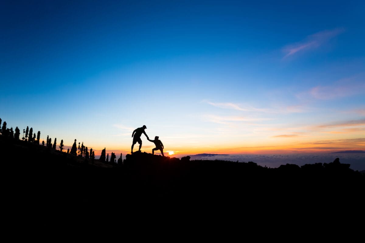 Two climbers help pull each other up in front of a sunset. 