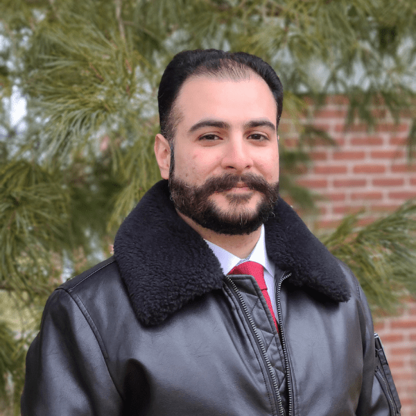 Ahmed Mohammed smiles at the camera in a black leather jacket in front of a pine tree.
