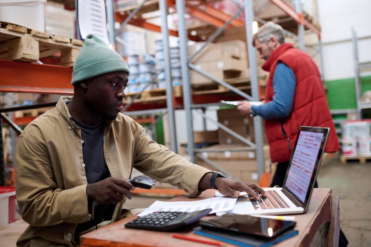 African American distributor using laptop to scan barcodes of parcels while sitting at his workplace at warehouse