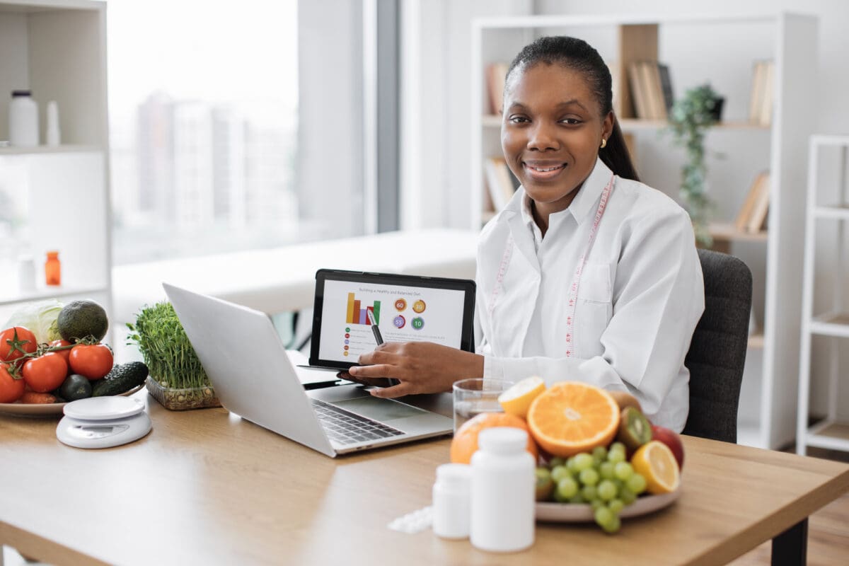 Portrait of smiling woman in lab coat holding digital tablet with diet plan while doing video conference via laptop. African american specialist in nutrition collaborate with health professionals.
