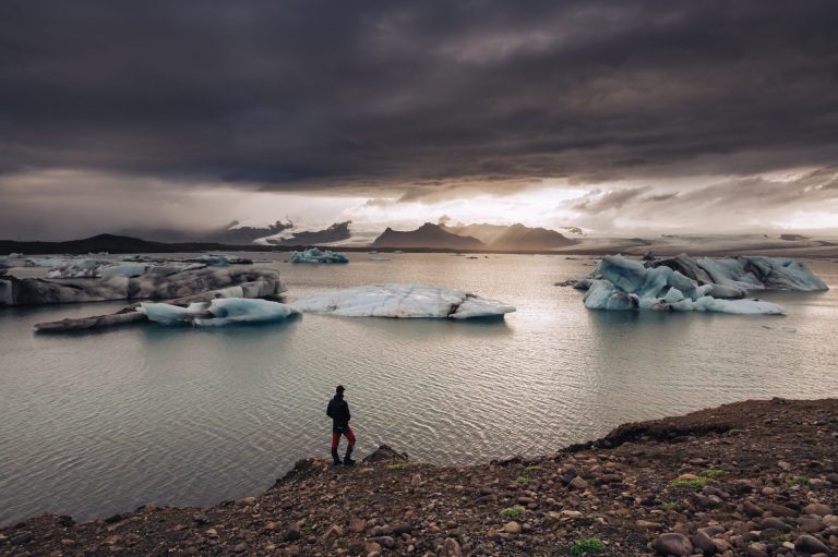 Man on a sea shore standing looking at water and icebergs.