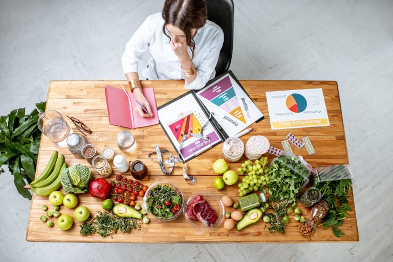 Woman writing a diet plan on a table full of healthy food options.