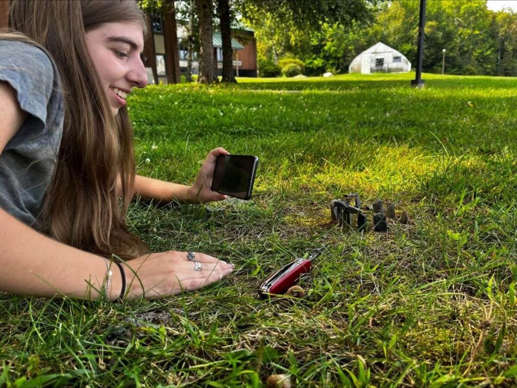 A younger person with a camera smiles at a tiny recreation of Stonehenge in the grass.
