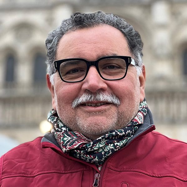 A man with black glasses and a red coat smiles at the camera.